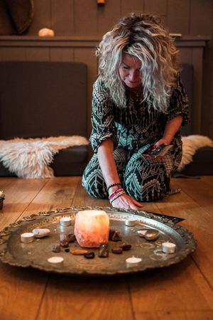 Person in patterned dress sits on floor with salt lamp, crystals and candles arranged on decorative metal tray.