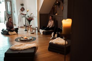 People sitting on meditation cushions in a cozy room with lit candles and natural light streaming through windows.