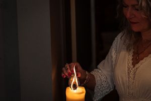 A person in a white lace sleeve lights a large yellow candle in a dark room, creating a warm glow.