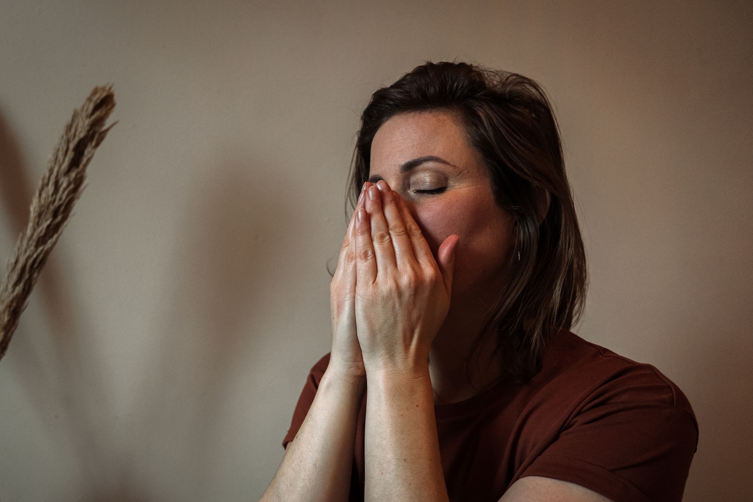 Person in brown shirt covering face with hands while praying or meditating against neutral wall background.