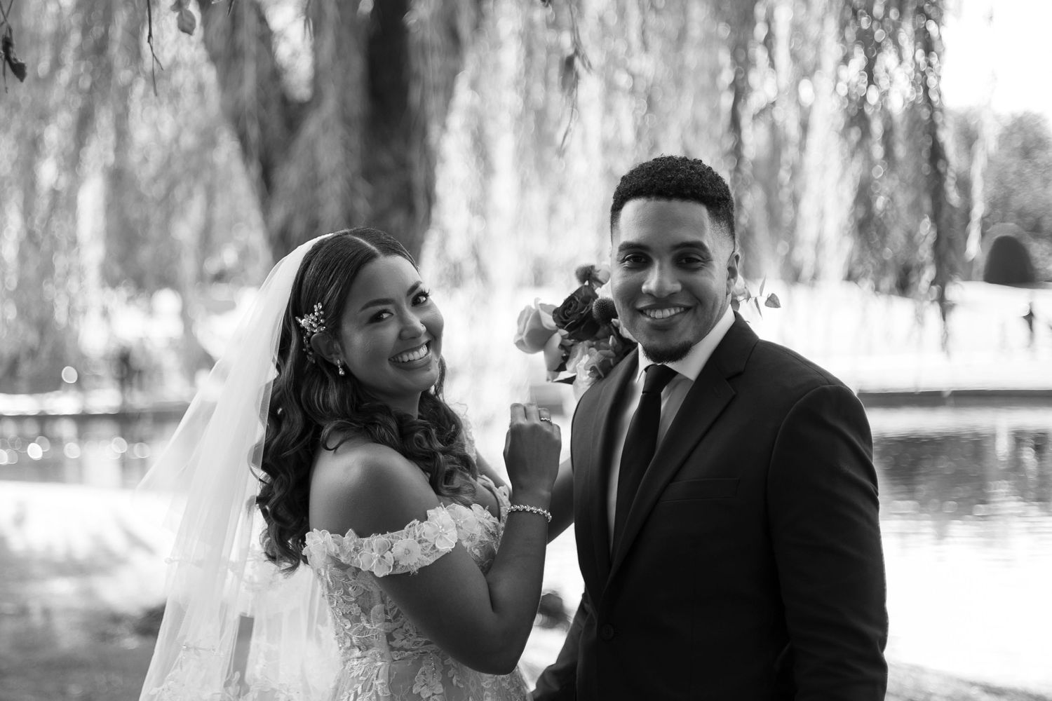 A bride and groom pose together outdoors near a willow tree and pond in a black and white bridal portrait.