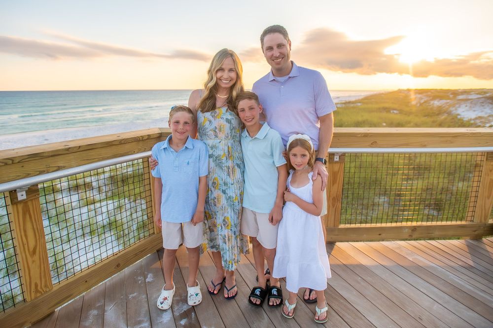 A family in coordinating white outfits poses for sunset photos on a wooden beach boardwalk.