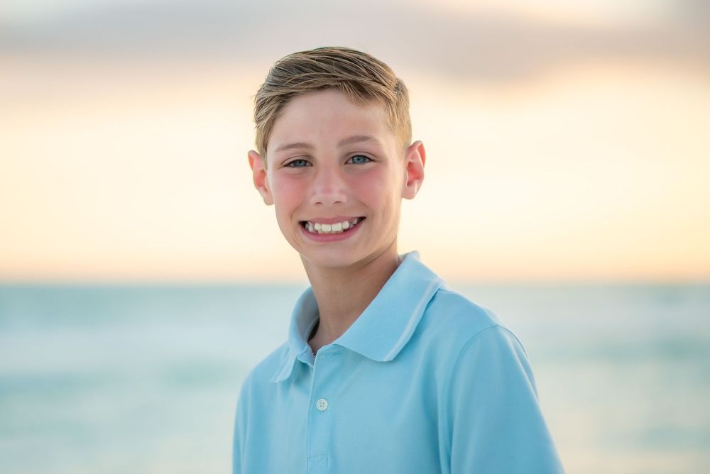 A young person in a light blue polo shirt smiles warmly against a soft sunset beach backdrop.