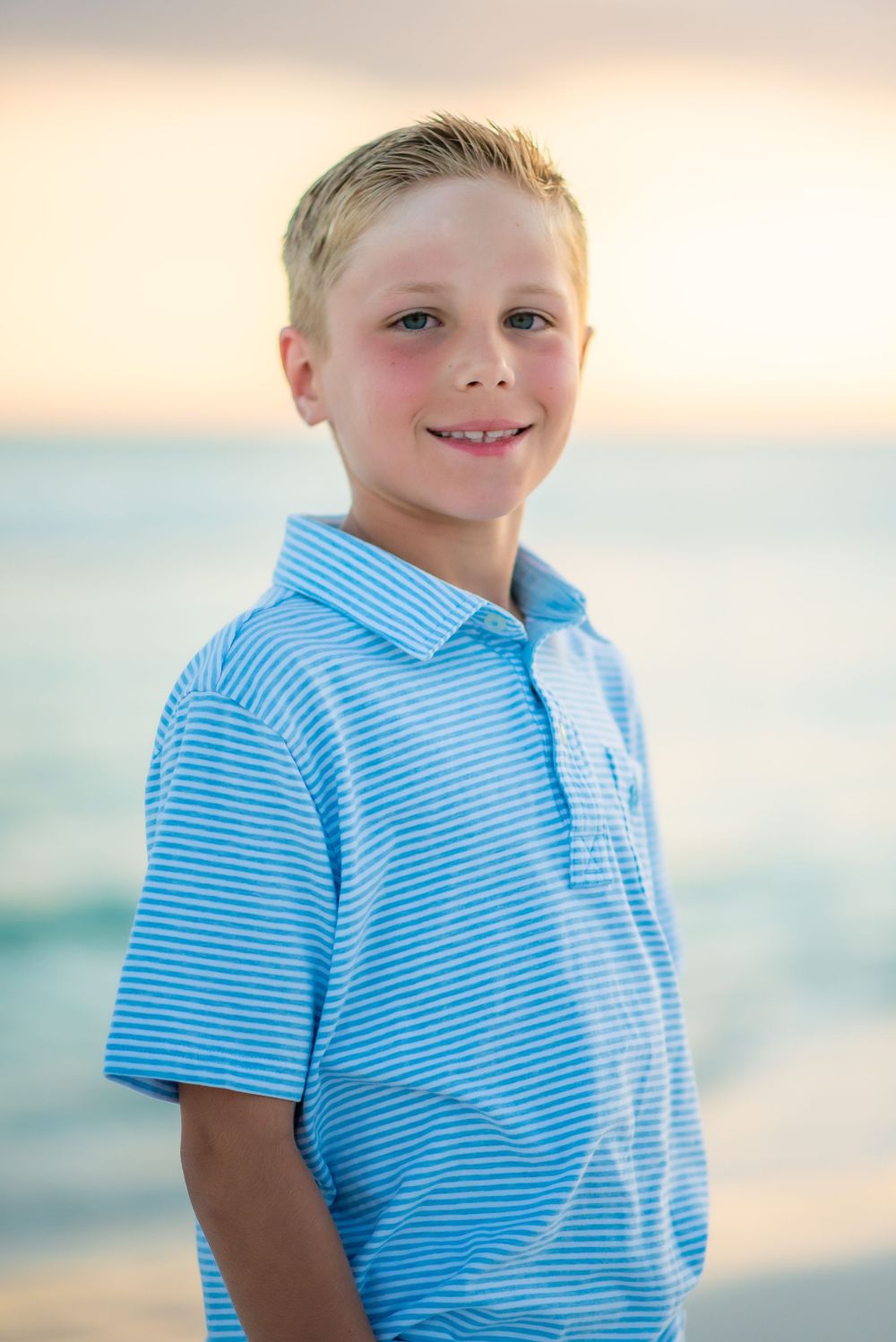 Child wearing blue and white striped polo shirt smiles at sunset on a beach.