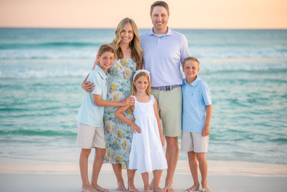 A family wearing coordinated blue and white outfits poses together on a sandy beach at sunset.