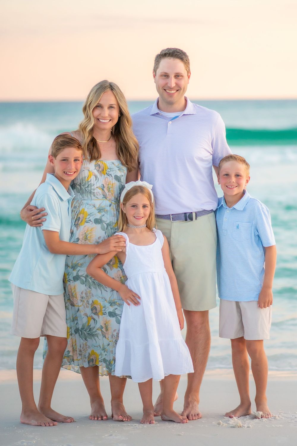A group in coordinated blue and white outfits poses together on a sandy beach with turquoise ocean waves in the background.