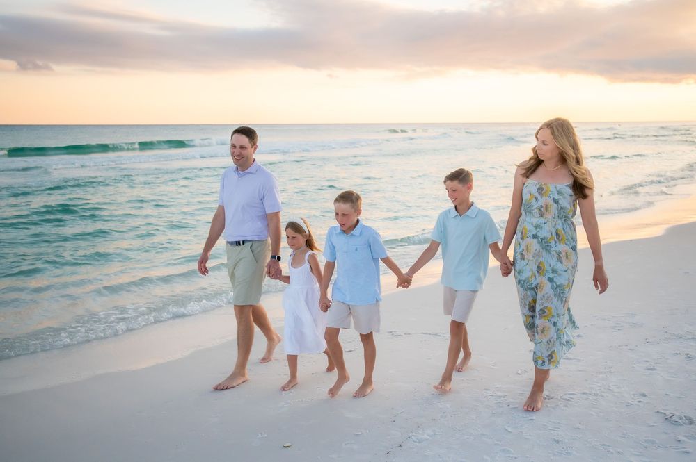 Family wearing white clothing walks together holding hands along a beautiful beach at sunset.