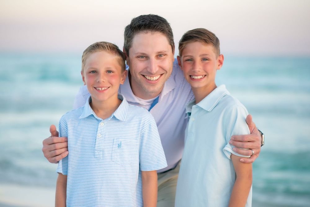 A family wearing coordinated blue and white outfits poses together on a sunny beach with ocean waves in the background.