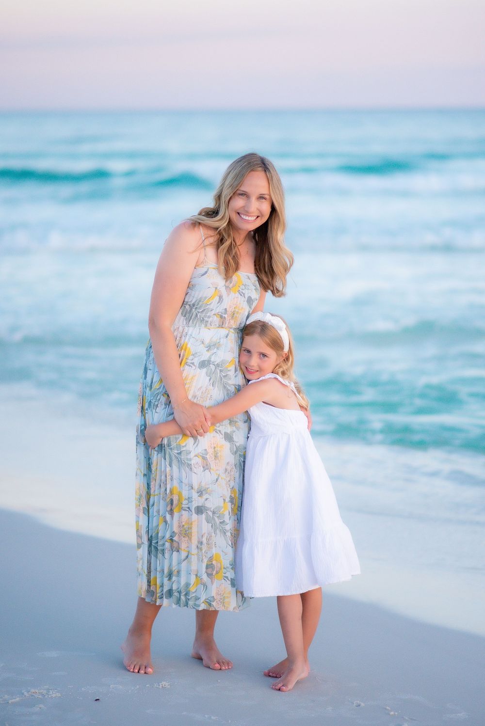 Mother and daughter share a tender embrace on a beach at sunset with turquoise waves and soft pink sky in the background.
