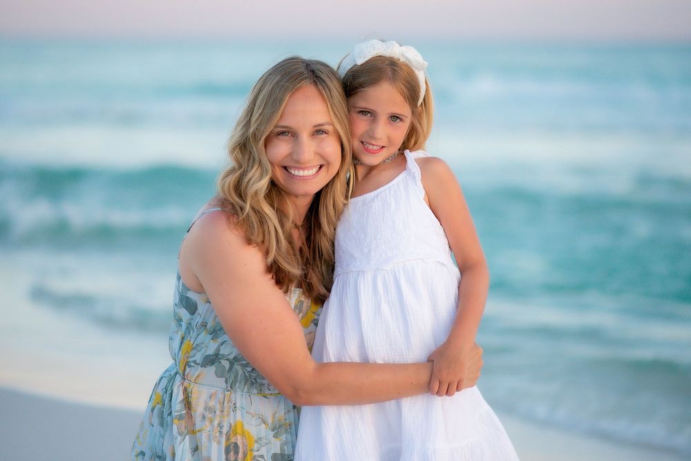 A loving family embraces while standing on a white sandy beach with gentle waves and pastel sky behind them.