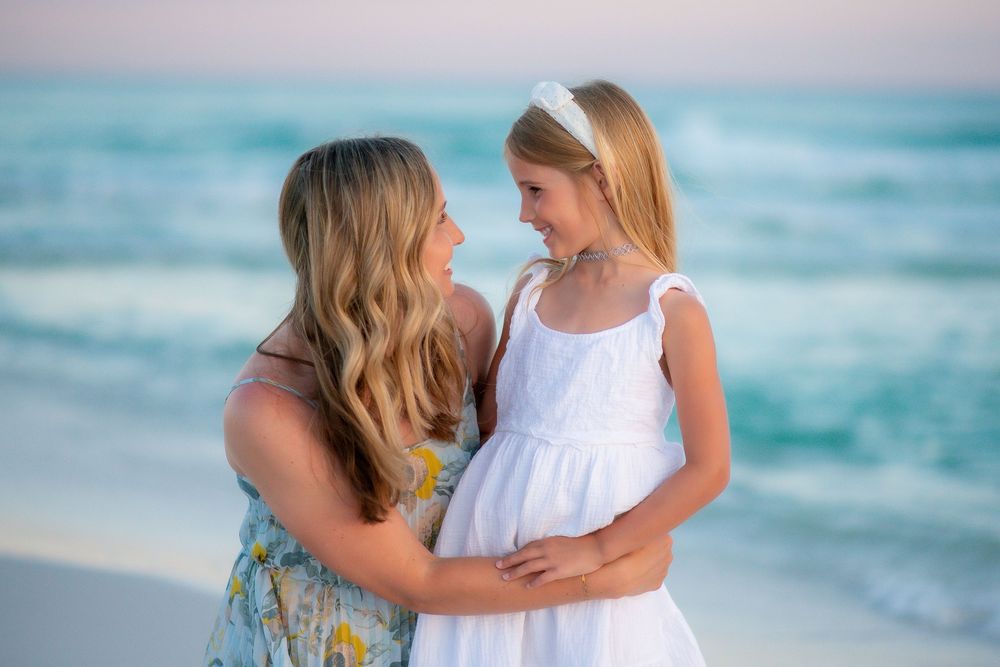 Two people in light summer dresses share a tender moment on a beach with turquoise ocean waves in the background.