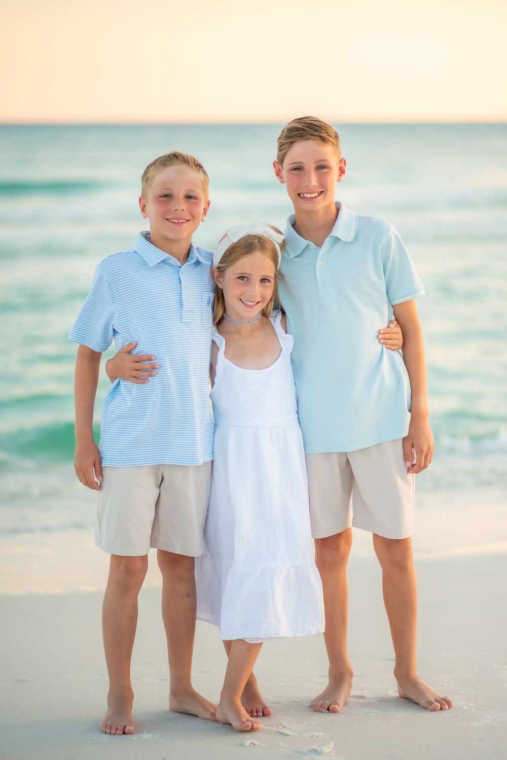 Three siblings in coordinating blue and white outfits pose together on a sunny beach with turquoise ocean waves.
