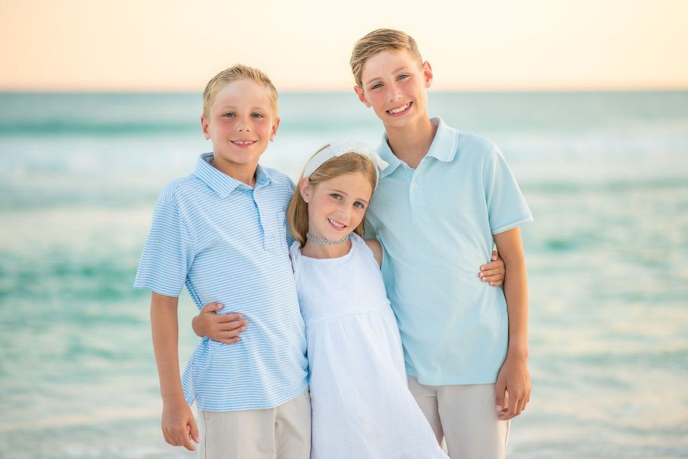 Family wearing coordinated blue and white outfits poses together on beach at sunset.