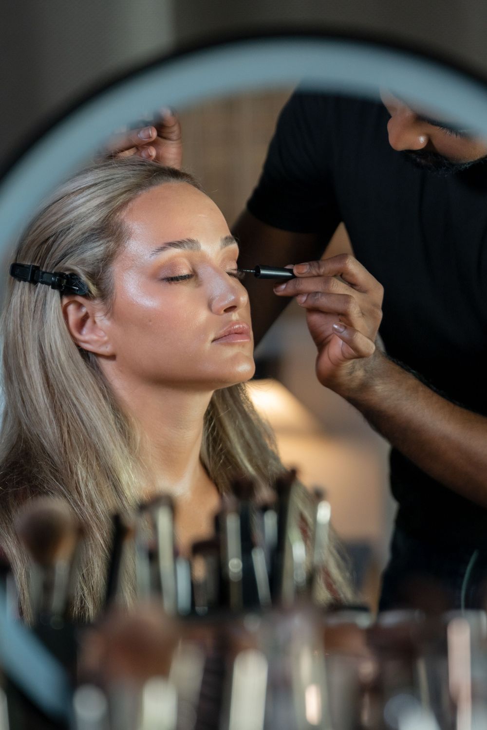 Professional makeup artist applying cosmetics to model's face in beauty salon with soft lighting and mirror reflections.