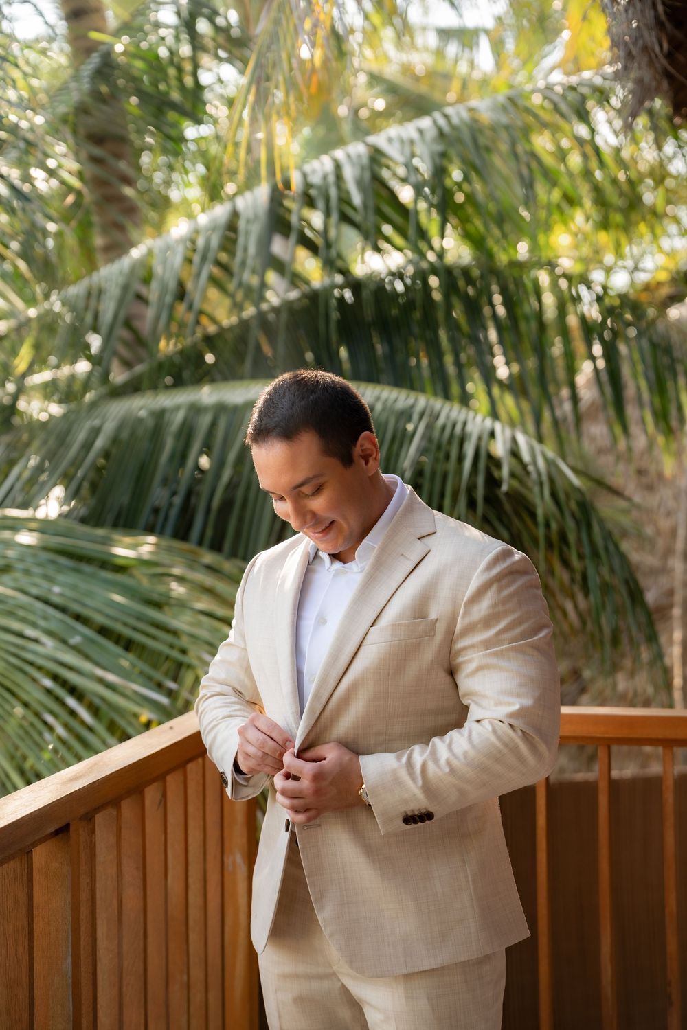 A groom in a light beige suit adjusts his boutonniere against a backdrop of tropical palm trees.