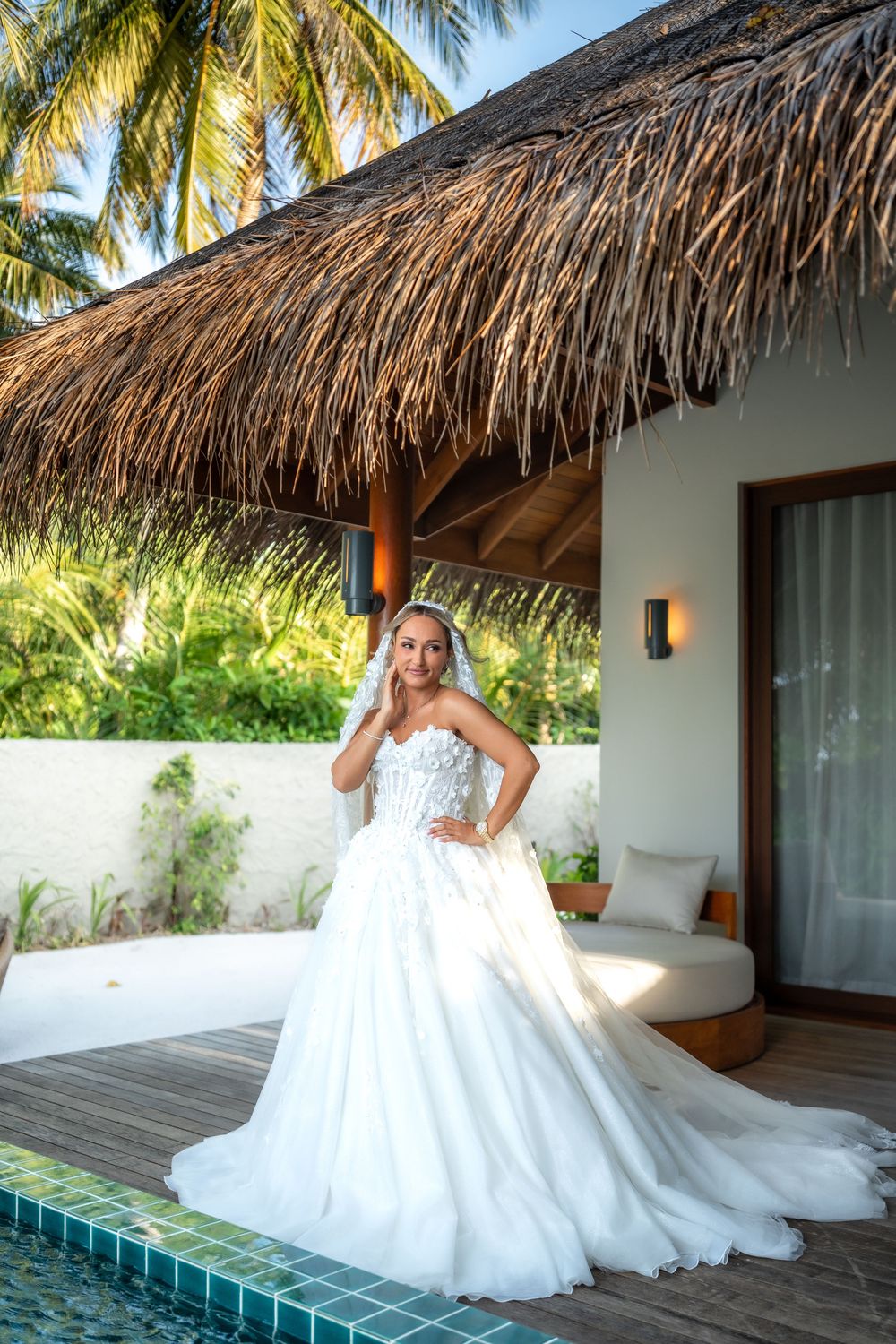 A white wedding dress flows elegantly across a tropical villa deck with palm trees and thatched roof in the background.