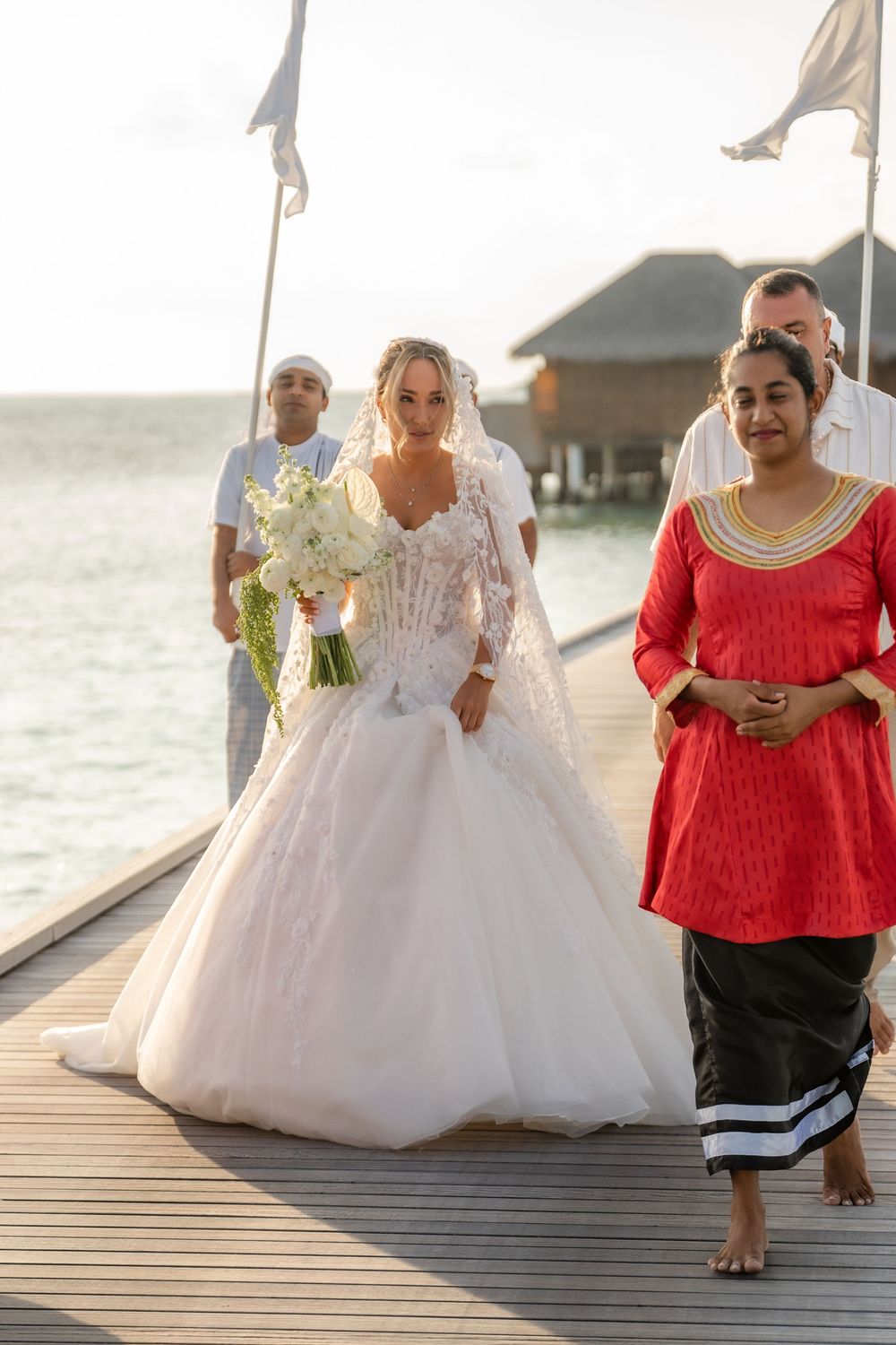 Wedding party walks along a wooden boardwalk over water at a tropical resort with a bride in a white gown.