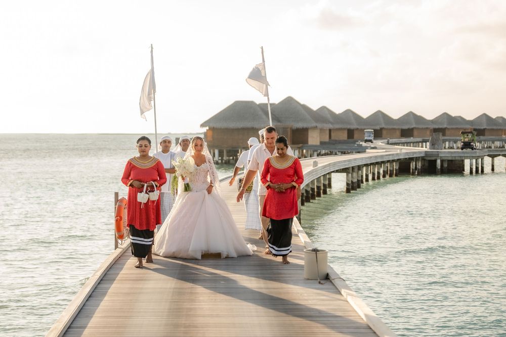 Wedding party in red and white attire poses on a wooden pier overlooking the ocean at sunset.