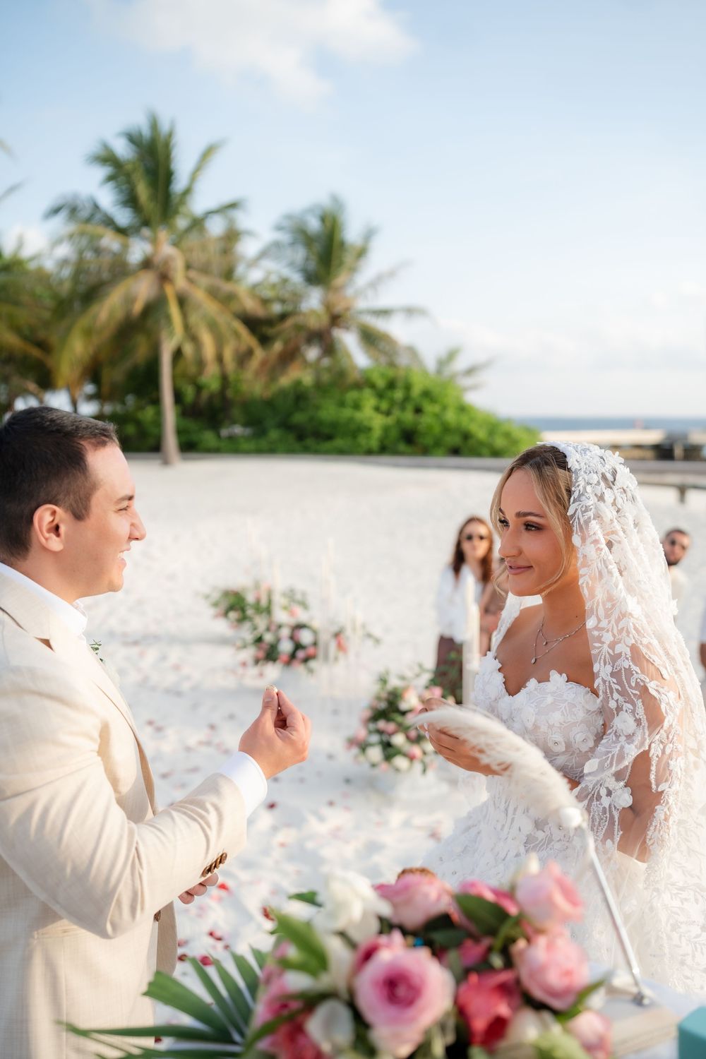 A couple shares an intimate moment during their beach wedding ceremony surrounded by pink flowers and palm trees.