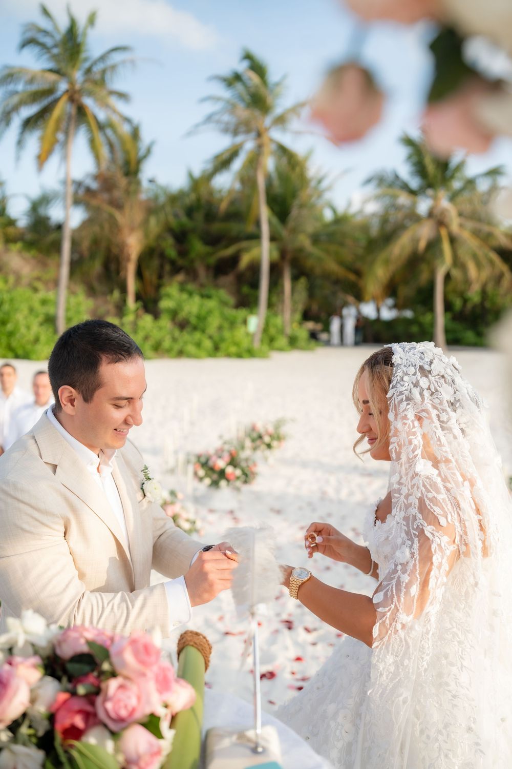 A couple shares an intimate moment under palm trees during a beach wedding ceremony with flowing white veil.