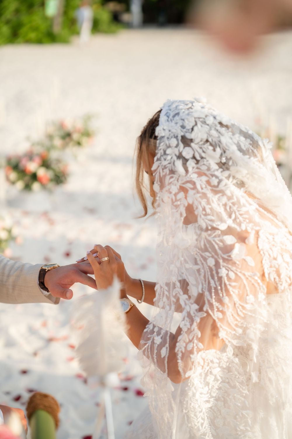 A romantic beach wedding moment with delicate lace veil and rose petals scattered on white sand.