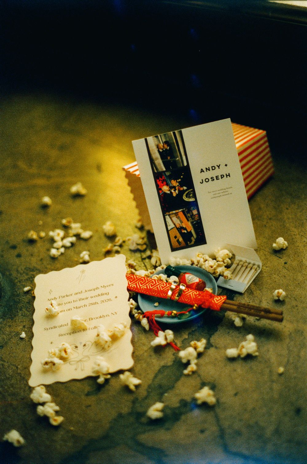 Wedding invitation for Andy and Joseph surrounded by scattered popcorn, a red party favor, and small spatula on a surface.