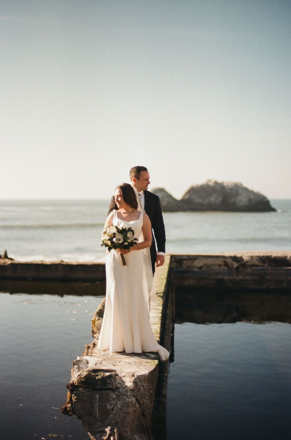Newlywed couple shares romantic moment on rocky coastal cliffs at sunset with ocean and mountains in background.