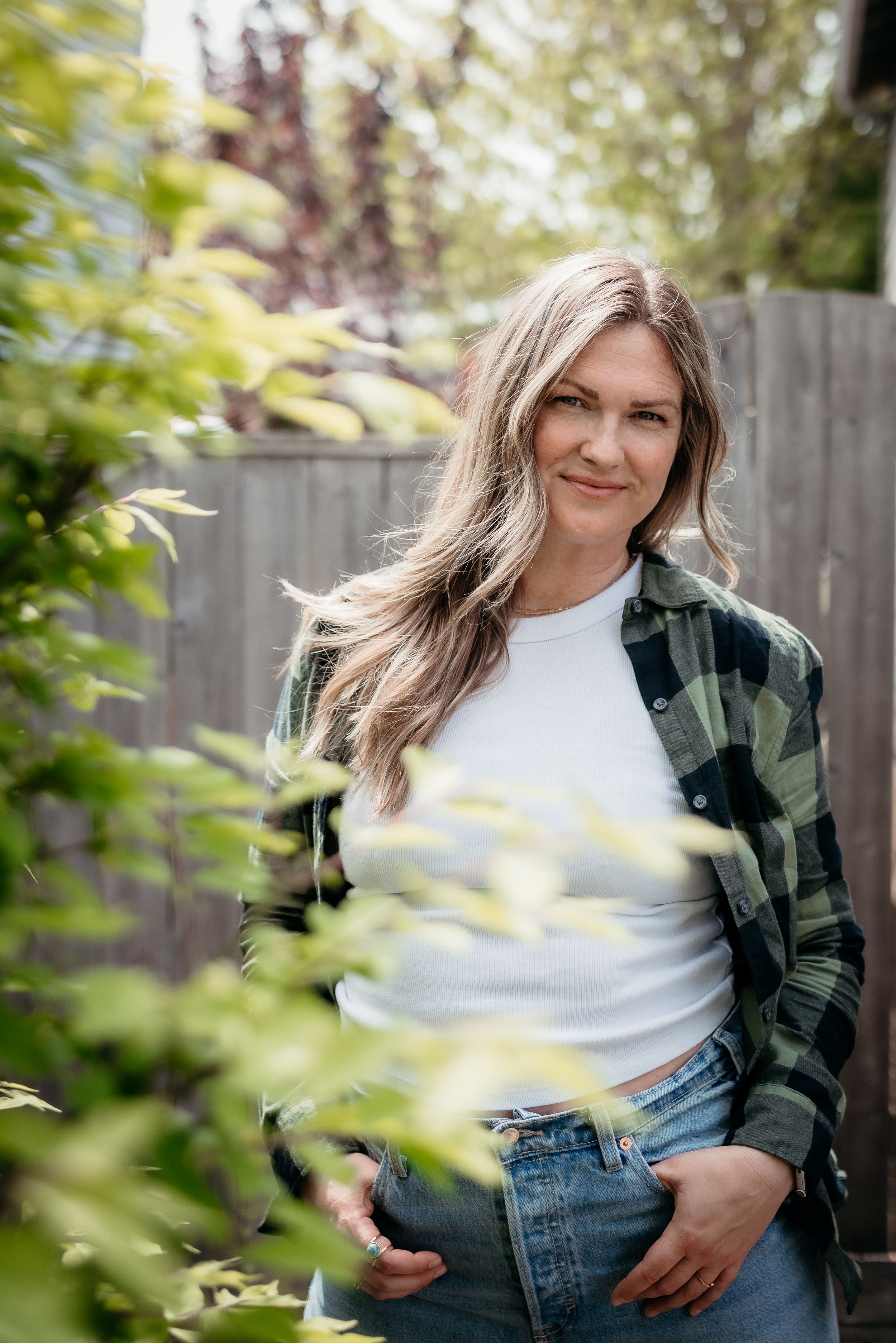 Woman with long blonde hair in plaid shirt and jeans smiling outdoors among green foliage.