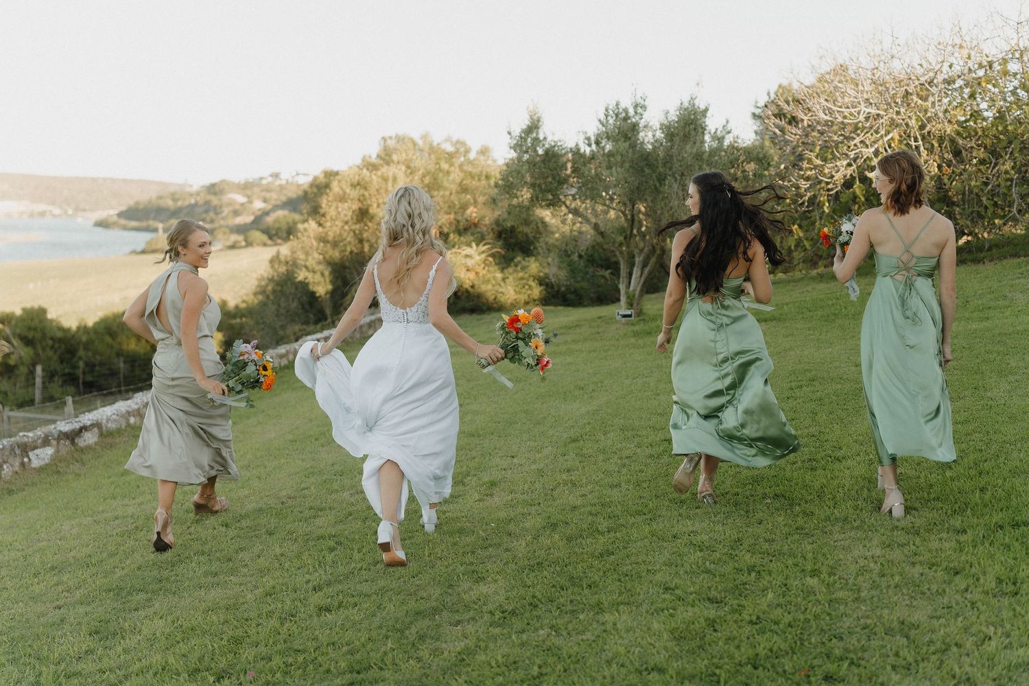 Wedding party running and dancing together on a grassy hillside in pastel dresses.