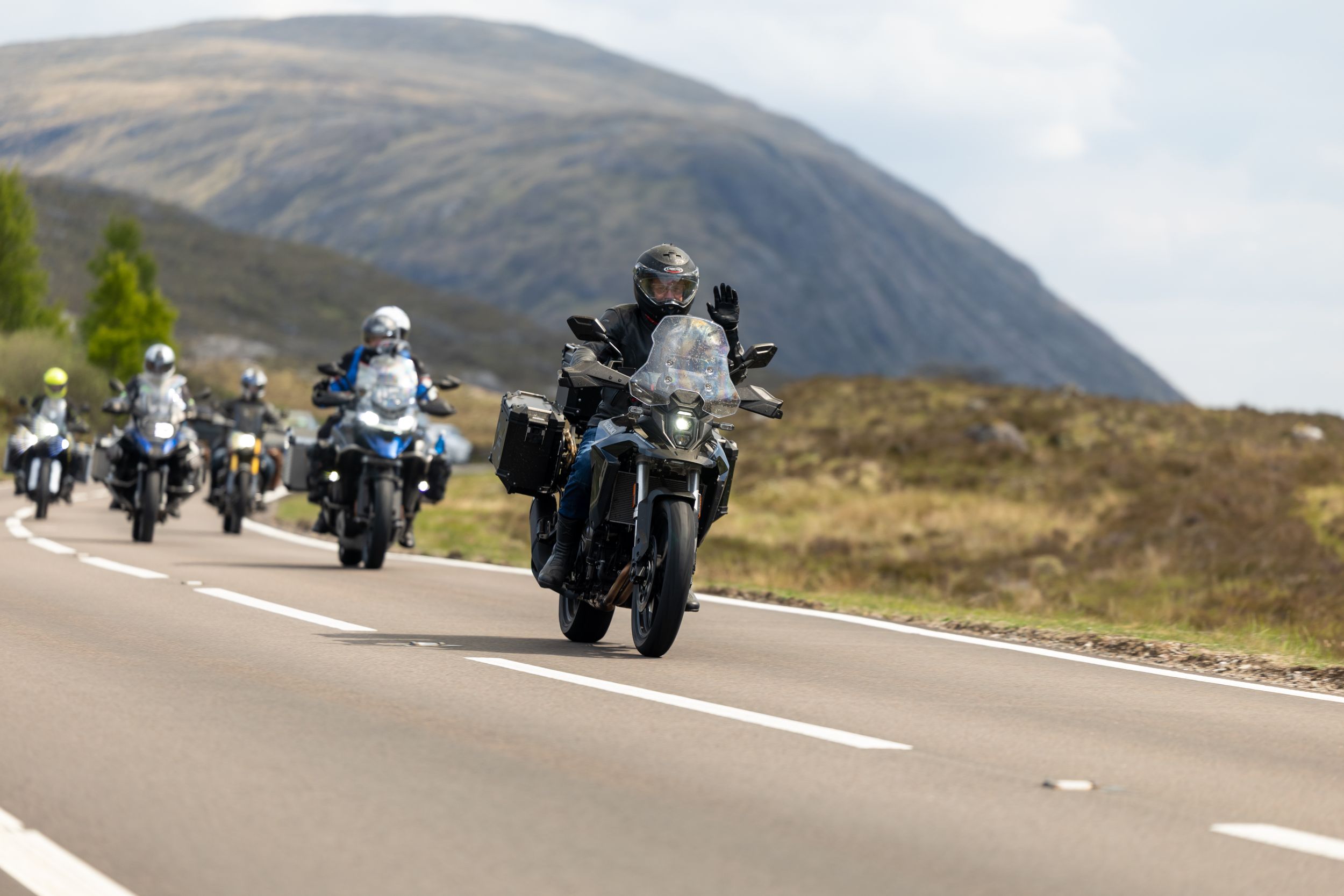 Group of motorcyclists riding in formation on a scenic mountain road with rolling hills in the background.