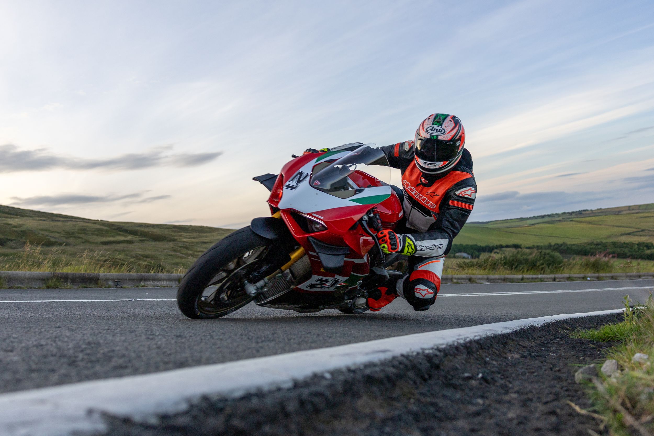 Motorcyclist in red racing gear leans into a curve while riding a sportbike on a scenic mountain road at dusk.