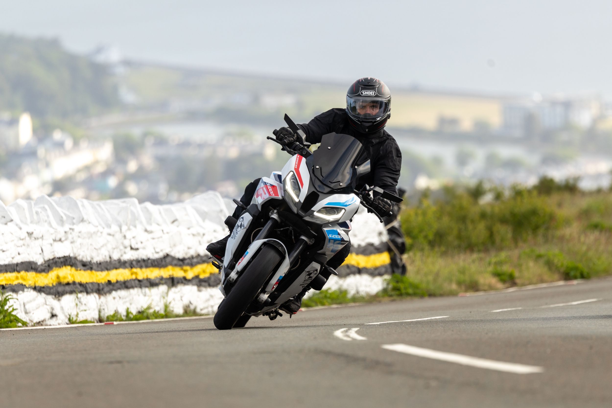A motorcyclist rides a black sport bike along a coastal road with white cliffs and ocean views in the background.