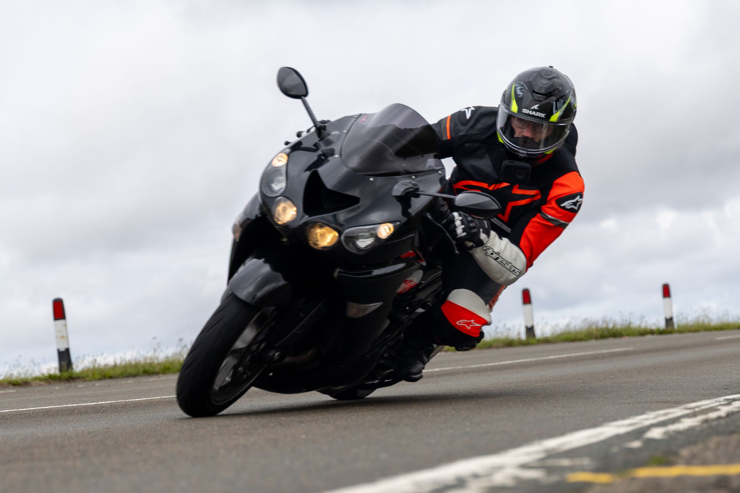 A motorcyclist in red and black gear leans into a turn while riding a black sport bike on a winding road.