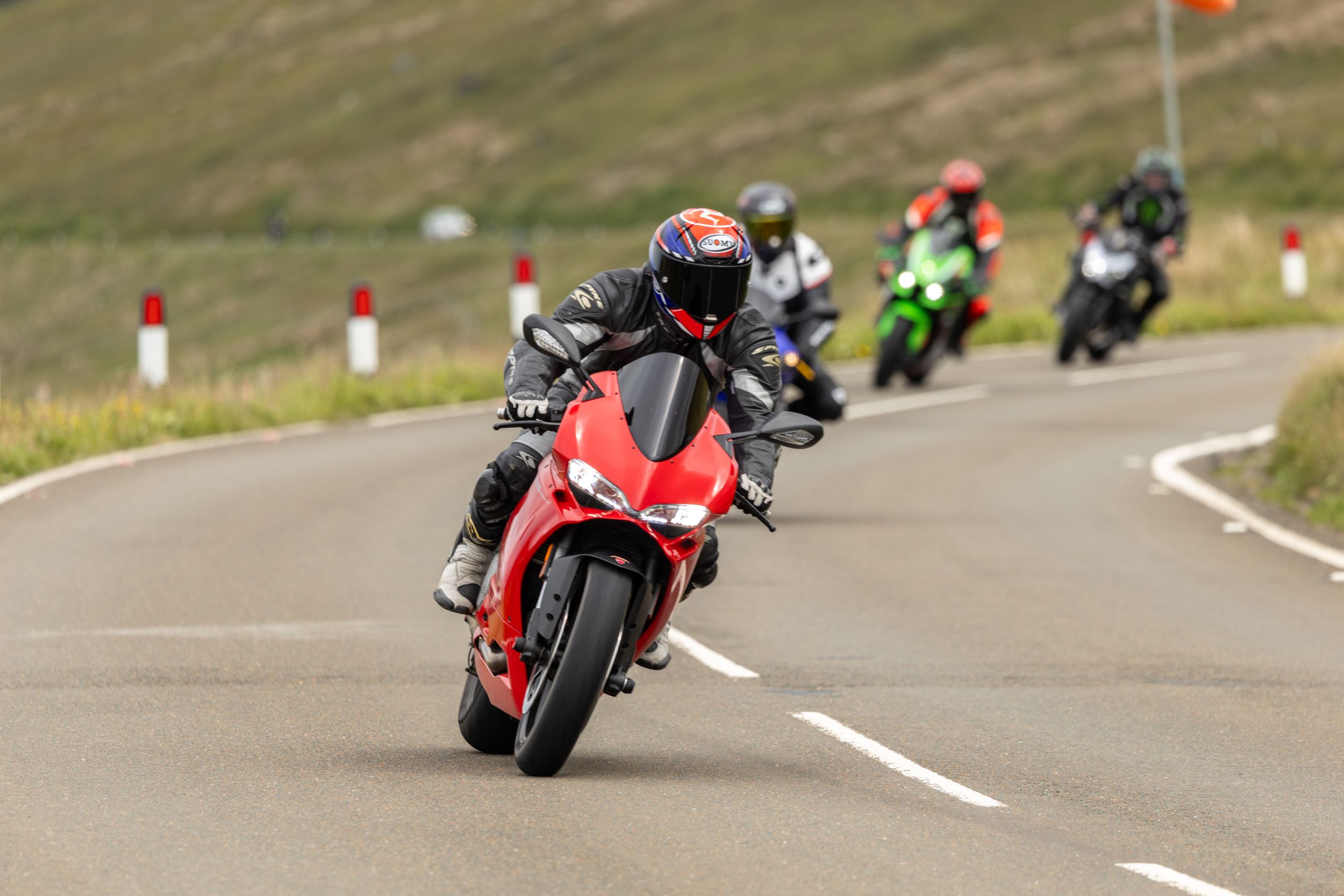 Motorcyclists racing on a winding mountain road with a red sport bike leading the pack.