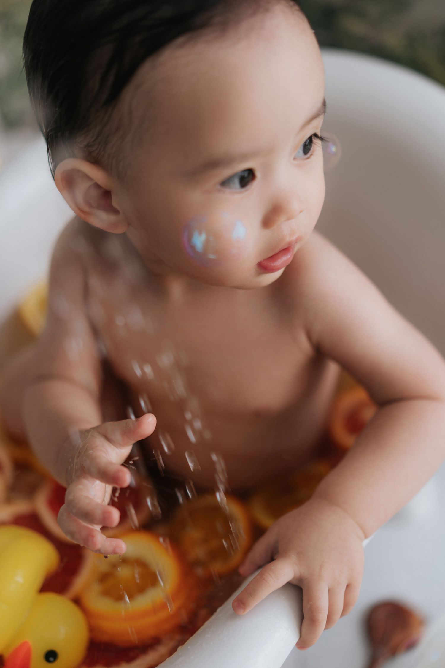 A baby in a high chair reaches for a rubber duck toy while splashing in water during bath or playtime.