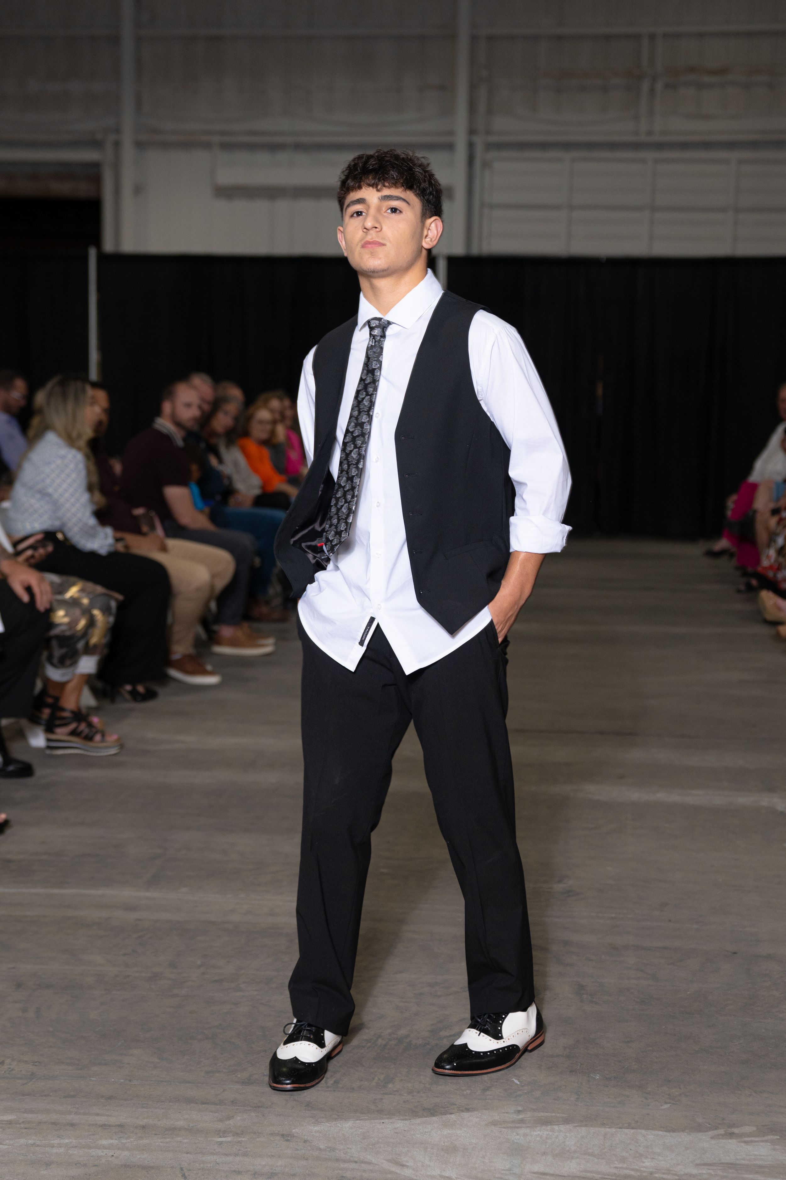 A model walks the runway wearing a formal black vest and pants with white dress shirt and black and white oxford shoes.