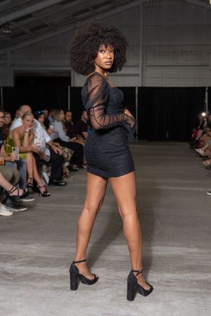 Model walks runway in black outfit and heels during fashion show with audience seated in background.