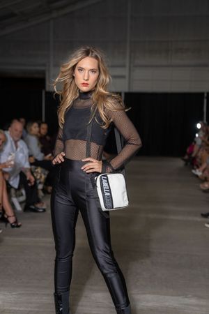 Model showcases edgy black outfit with mesh top, leather pants and white handbag on industrial runway.