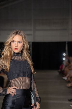 Fashion model walks runway wearing black mesh top and dark ensemble during fashion show presentation.