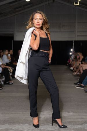 Model poses confidently on runway wearing black pants, crop top and white blazer with high heels.