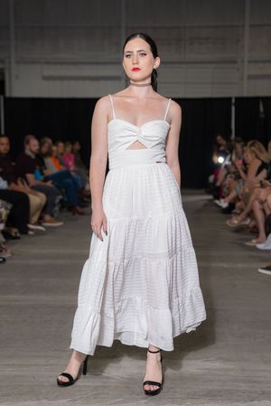 Model walks runway in white tiered maxi dress with cutout detail and black sandals during fashion show.