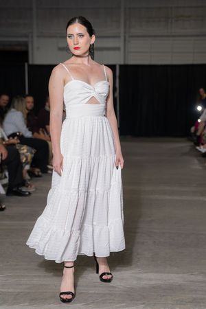 Model walks runway in white tiered maxi dress with cutout detail and black sandals at fashion show.