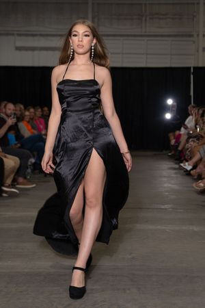 Model struts in flowing black satin evening gown on industrial runway with audience visible in background.