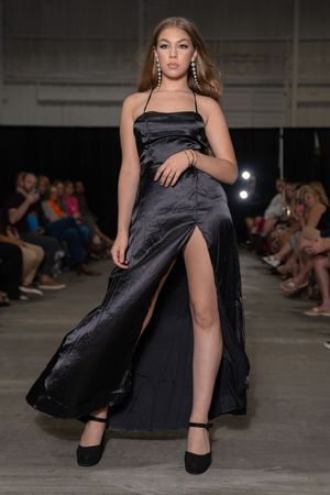 Model showcases elegant black satin evening gown with thigh-high slit on industrial runway.