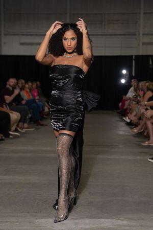 Model in black crop top and sheer stockings walks runway during fashion show with audience seated in background.