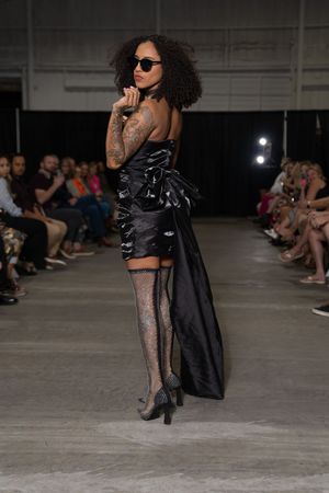 Fashion model walks runway in black evening wear and patterned stockings at an industrial-style fashion show.