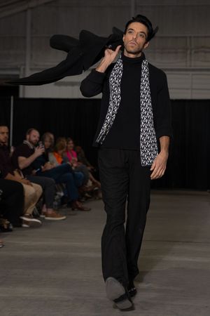 Fashion model in black outfit and patterned scarf performs dynamic dance moves on runway during fashion show.