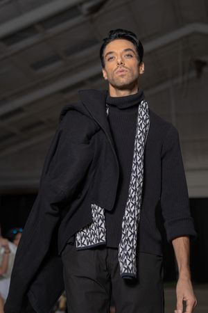 Model showcases black winter coat with geometric patterned scarf and turtleneck on industrial runway.