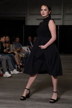 Model in sleeveless black dress and platform sandals walks down concrete runway at fashion show.