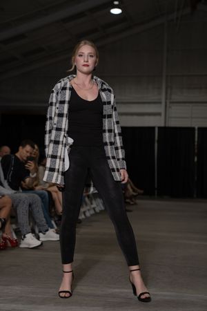 Model showcases black outfit with plaid jacket on industrial-style runway with audience seated along sides.
